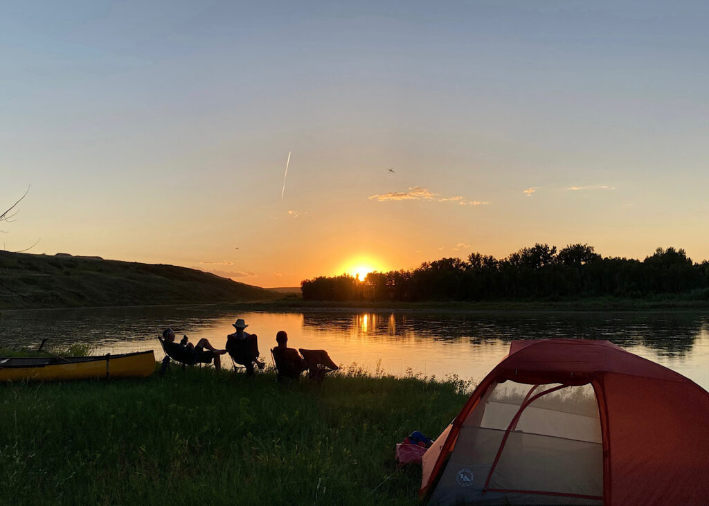 Sunset above river with tent in foreground and people sitting on riverbank.