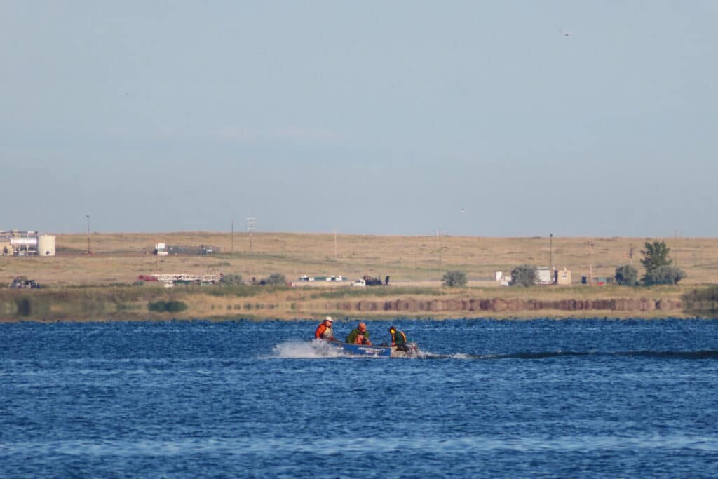 Three men in aluminum boat heading out across lake with prairie grasslands in distance.